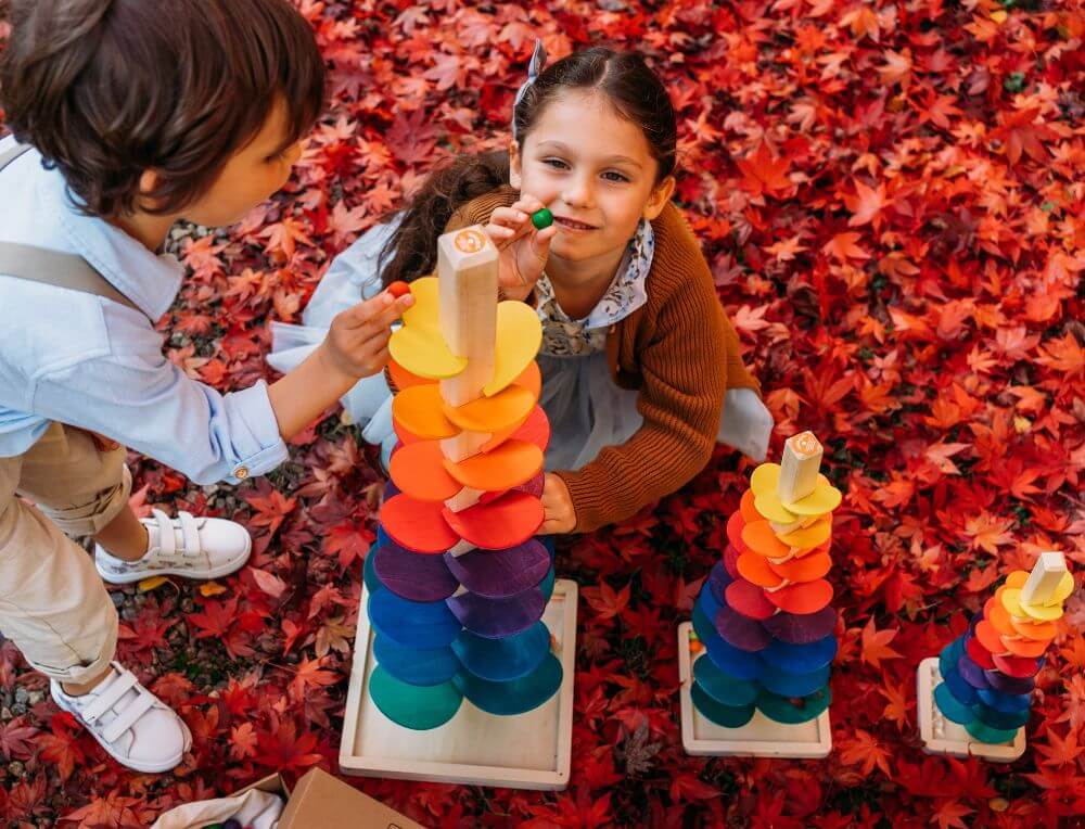 Children placing wooden marbles on a wooden marble tree toy - the largest of three - in an outdoor setting with Autumn leaves